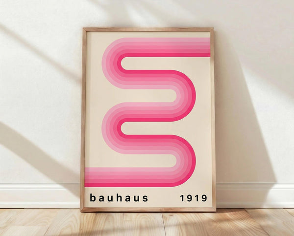 May include: Pink gradient ribbon forming wavy S-shaped curves on cream background, displayed in thin wood frame leaning against white textured wall on light wooden floor. Text reads "bauhaus 1919" in black lowercase letters at bottom. Soft natural lighting creates gentle shadows on the wall.