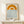 May include: Framed geometric art print featuring orange semicircular sun design above blue wavy lines representing water, with "bauhaus 1919" text at bottom. Print displayed in natural wood frame on light wooden shelf next to small white wrapped gift with evergreen sprig. Clean white wall background with soft natural lighting.