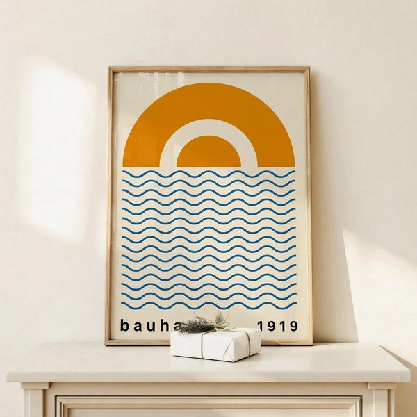 May include: Framed geometric art print featuring orange semicircular sun design above blue wavy lines representing water, with "bauhaus 1919" text at bottom. Print displayed in natural wood frame on light wooden shelf next to small white wrapped gift with evergreen sprig. Clean white wall background with soft natural lighting.