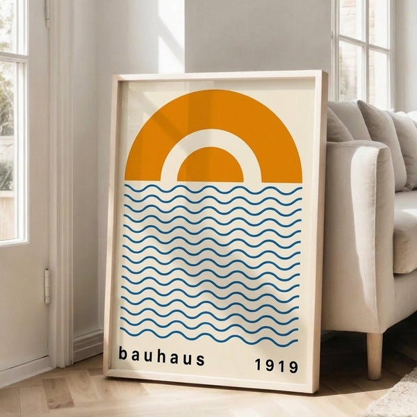 May include: Framed poster with cream background featuring orange and white concentric semicircles above blue wavy horizontal lines, text reading "bauhaus 1919" below. Light wood frame leans against white wall next to beige upholstered sofa in bright room with large windows and natural light streaming across wooden floors.
