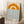 May include: Framed poster with minimalist design featuring orange and cream concentric arcs resembling a sun above horizontal blue wavy lines representing water. Text reads "bauhaus 1919" in black lowercase letters. Light wood frame leans against white wall corner next to wooden block with white coffee cup and saucer, on white textured rug with beige fabric visible.