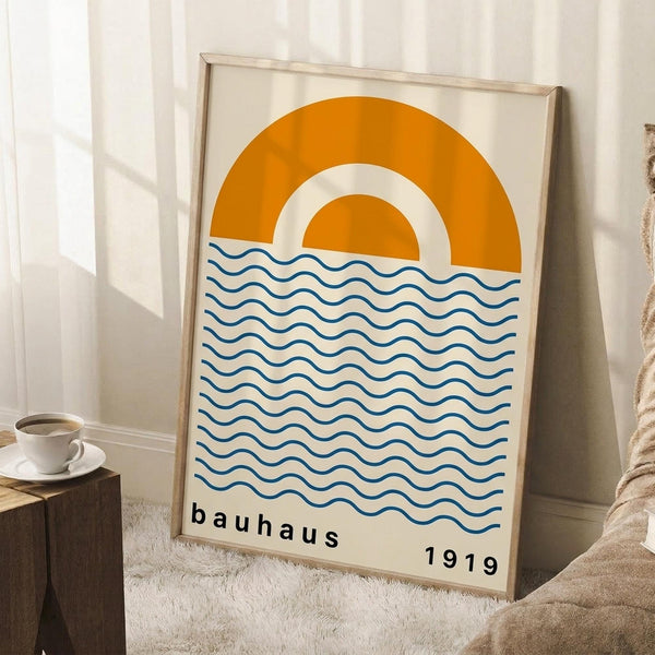 May include: Framed poster with minimalist design featuring orange and cream concentric arcs resembling a sun above horizontal blue wavy lines representing water. Text reads "bauhaus 1919" in black lowercase letters. Light wood frame leans against white wall corner next to wooden block with white coffee cup and saucer, on white textured rug with beige fabric visible.