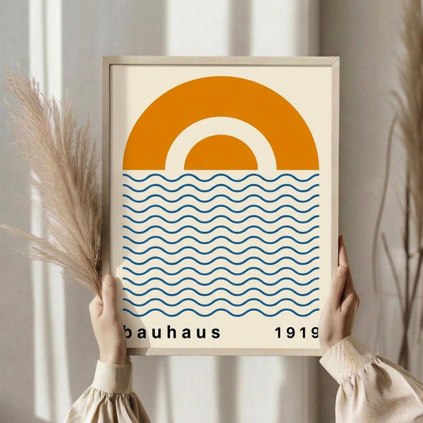 May include: Person in beige ruffled sleeves holding framed minimalist poster featuring orange semicircular sun design above blue wavy lines representing water, with "bauhaus 1919" text below. Light wooden frame against neutral curtained background with dried pampas grass visible on left side.