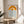 May include: A framed minimalist poster with an orange gradient rainbow arc above wavy blue lines representing water, displayed on a light wood round side table next to a beige upholstered chair. Natural sunlight streams across the white wall creating soft shadows from a plant visible in the corner.