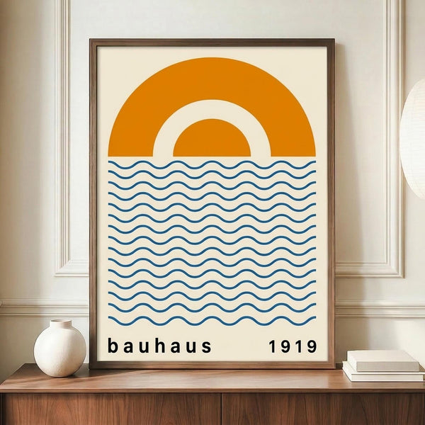 May include: A framed minimalist poster featuring an orange semicircle sun above blue wavy lines representing water, with "bauhaus 1919" text below. The artwork is displayed in a thin brown frame on a wooden shelf next to a small cream ceramic vase and white books against beige paneled walls.