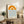 May include: Framed minimalist print with orange geometric sun design and blue wavy lines below, displayed on wooden sideboard beside fern plant, ceramic vase, and books in bright living room with beige chair