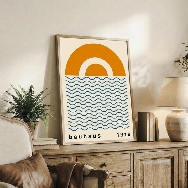 May include: Framed minimalist print with orange geometric sun design and blue wavy lines below, displayed on wooden sideboard beside fern plant, ceramic vase, and books in bright living room with beige chair