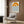 May include: Framed poster with orange concentric circles resembling a sun above blue wavy lines representing water, displayed on wood-paneled wall above beige upholstered chair with geometric patterned pillow. Modern minimalist room setting with brass floor lamp and dark round side table with open book.