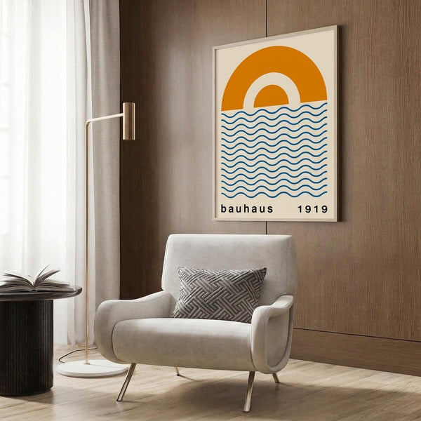 May include: Framed poster with orange concentric circles resembling a sun above blue wavy lines representing water, displayed on wood-paneled wall above beige upholstered chair with geometric patterned pillow. Modern minimalist room setting with brass floor lamp and dark round side table with open book.