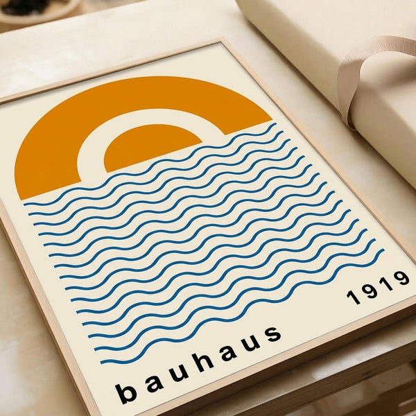 May include: Framed minimalist poster with orange semicircular sun and blue wavy lines representing water, displayed on cream colored surface next to rolled papers. The design features "bauhaus" and "1919" text in black lowercase letters on an off-white background within a thin wooden frame.
