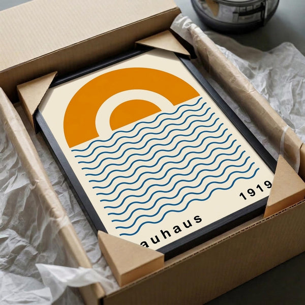 May include: Framed minimalist art print in black frame, packed in cardboard box with tissue paper. Design features orange semicircular sun and blue wavy lines representing water on cream background, with "1919" and "bauhaus" text at bottom. Wooden dowels and packaging materials visible around shipping box.