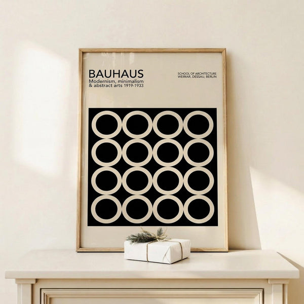May include: Framed Bauhaus modernist poster with black background and white circle pattern arranged in grid, displayed on light wood console table beside small wrapped gift with greenery, against cream wall in minimalist room setting