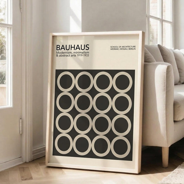 May include: Framed Bauhaus poster with black background and cream-colored circles arranged in a 4x4 grid pattern, leaning against white wall next to beige upholstered chair. Poster features "BAUHAUS" text and "Modernism, minimalism & abstract arts 1919-1933" subtitle. Natural light streams through white-framed windows onto light wooden floors in minimalist living room setting.