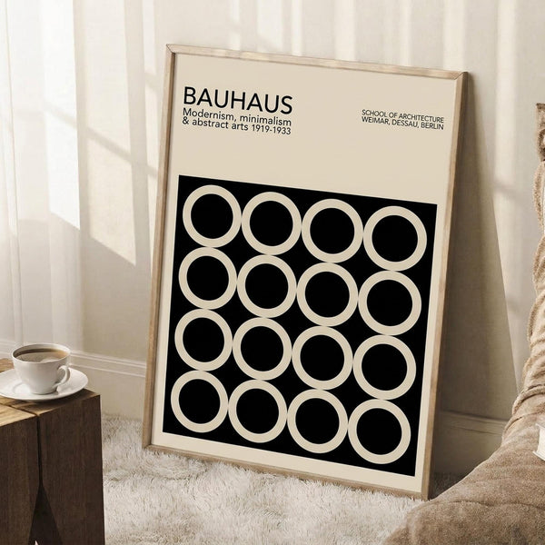 May include: Framed Bauhaus poster with black background and white circles arranged in geometric grid pattern, leaning against white wall corner. Light wood frame, cream matting, minimalist typography at top. Wooden block with white coffee cup nearby on textured rug in bright, airy room setting.