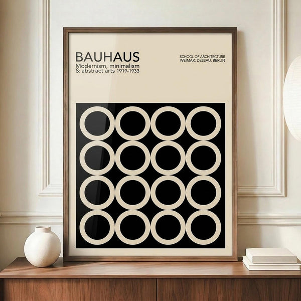 May include: Framed Bauhaus poster with black background and grid of white circles in various styles, displayed on wooden sideboard next to white ceramic vase and books. Cream colored walls with white trim molding visible in background. Modern minimalist interior styling with warm wood tones.