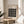 May include: Framed Bauhaus poster with black and white geometric circle pattern displayed on wooden round side table next to beige upholstered chair. Natural sunlight casts shadows across neutral-toned room with plant leaves visible in corner. Poster features modernist design with grid of white circles on black background in light wood frame.