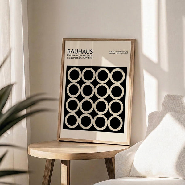 May include: Framed Bauhaus poster with black and white geometric circle pattern displayed on wooden round side table next to beige upholstered chair. Natural sunlight casts shadows across neutral-toned room with plant leaves visible in corner. Poster features modernist design with grid of white circles on black background in light wood frame.