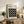 May include: Framed Bauhaus poster with black background and white circles arranged in a 4x4 grid pattern, displayed on rustic wooden sideboard alongside potted fern, ceramic vase, and stacked books in neutral-toned living room with beige upholstered chair and cream walls