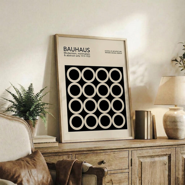 May include: Framed Bauhaus poster with black background and white circles arranged in a 4x4 grid pattern, displayed on rustic wooden sideboard alongside potted fern, ceramic vase, and stacked books in neutral-toned living room with beige upholstered chair and cream walls