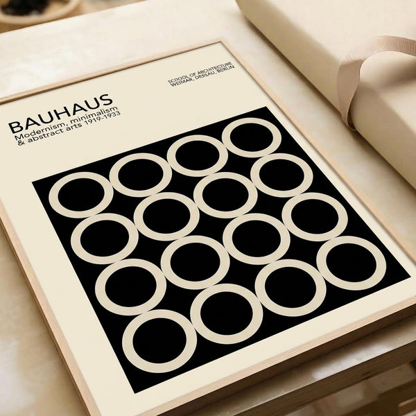 Minimalist Bauhaus-inspired poster with geometric pattern of white circles on black background, displayed in cream-colored frame on neutral textured surface alongside rolled paper tubes. Clean modernist design featuring repeated circular motifs in monochromatic color scheme, photographed at an angle showing the artwork's crisp geometric forms and gallery-style presentation.