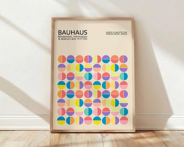May include: A framed poster featuring colorful geometric circles in a grid pattern with "BAUHAUS" text at the top, displayed in a light wooden frame against a white wall on wooden flooring with natural window light casting shadows across the scene.