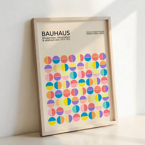 May include: Framed Bauhaus poster with colorful geometric circles in various bright colors including yellow, pink, blue, orange, and purple arranged in a grid pattern. The cream-colored print features "BAUHAUS" text at top with "Modernism, minimalism & abstract arts 1919-1933" and "School of Architecture Weimar, Dessau, Berlin" below. Light wood frame leans against white wall creating soft shadows.