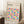 May include: Framed Bauhaus poster with colorful geometric circles in pink, blue, yellow, orange, and purple tones leaning against white wall corner. Light wood frame, cream background with modernist typography. Coffee cup on wooden block nearby, beige throw blanket visible, natural lighting creates soft shadows on white textured rug.