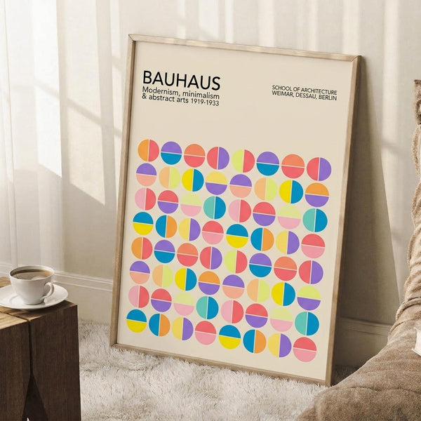 May include: Framed Bauhaus poster with colorful geometric circles in pink, blue, yellow, orange, and purple tones leaning against white wall corner. Light wood frame, cream background with modernist typography. Coffee cup on wooden block nearby, beige throw blanket visible, natural lighting creates soft shadows on white textured rug.