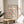 May include: Framed Bauhaus poster with colorful geometric circles in yellow, pink, blue, orange, and purple displayed on light wood round side table next to beige upholstered chair. Natural sunlight streams across white walls creating soft shadows. Green plant leaves visible in corner. Modern minimalist room setting with neutral tones.