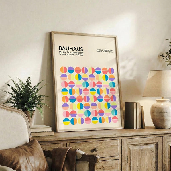 May include: Framed Bauhaus poster with colorful geometric circles in bright blues, yellows, pinks, and oranges displayed on rustic wooden sideboard alongside fern plant, ceramic vase, and stacked books in neutral living room setting with beige walls and upholstered furniture.
