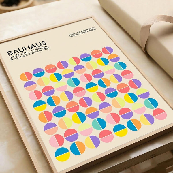 May include: Bauhaus-style poster print with colorful geometric circles arranged in a grid pattern, featuring vibrant blues, pinks, yellows, oranges, and purples on cream background. The framed artwork sits on a light wooden surface next to rolled paper tubes, showing modernist minimalist design with split-circle motifs throughout the composition.