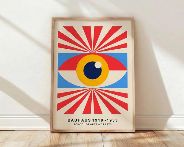 May include: Framed poster with geometric Bauhaus-style design featuring a large stylized eye with yellow iris, dark blue pupil, and white highlight, surrounded by blue rectangular background. Red and white radiating sunburst rays extend above and below the eye. Text at bottom reads "BAUHAUS 1919-1933 SCHOOL OF ARTS & CRAFTS". Natural wood frame sits on light wooden floor against white wall with soft shadows.