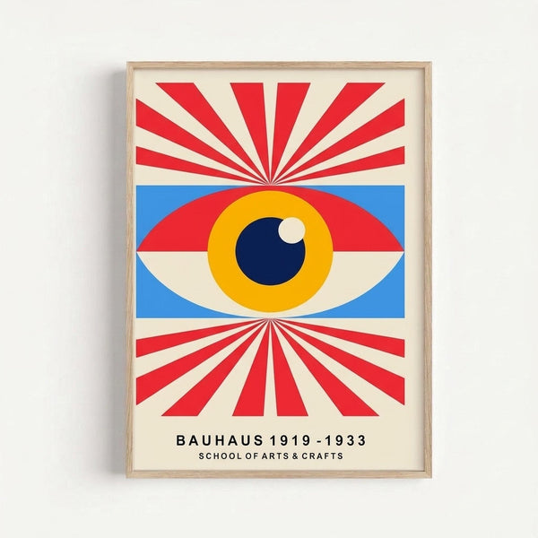Framed poster featuring a stylized eye with yellow iris and dark blue pupil centered on a blue background, surrounded by radiating red and cream sunburst rays extending to frame edges. Text below reads "BAUHAUS 1919-1933 SCHOOL OF ARTS & CRAFTS" in black letters. Light wood frame mounted on white wall.