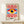 May include: Framed poster featuring a stylized eye with yellow iris, dark blue pupil, and white highlight, set against a blue rectangular background. Red and white radiating stripes extend above and below the eye from center points. Text reads "BAUHAUS 1919-1933 SCHOOL OF ARTS & CRAFTS" at bottom. Displayed in light wood frame on white wooden shelf next to small wrapped gift with twine and greenery sprig against cream wall.