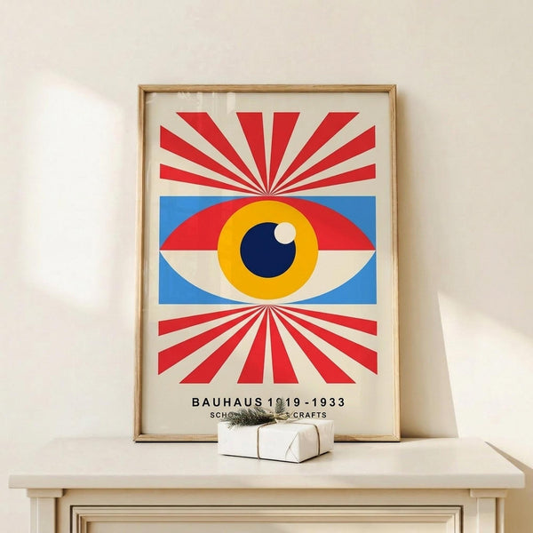 May include: Framed poster featuring a stylized eye with yellow iris, dark blue pupil, and white highlight, set against a blue rectangular background. Red and white radiating stripes extend above and below the eye from center points. Text reads "BAUHAUS 1919-1933 SCHOOL OF ARTS & CRAFTS" at bottom. Displayed in light wood frame on white wooden shelf next to small wrapped gift with twine and greenery sprig against cream wall.
