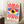 May include: Framed poster with abstract eye design featuring red and white radiating sunburst pattern, central eye with yellow iris and dark blue pupil surrounded by blue geometric shapes, displayed leaning against white wall next to wooden side table with white coffee cup and cozy knit throw blanket