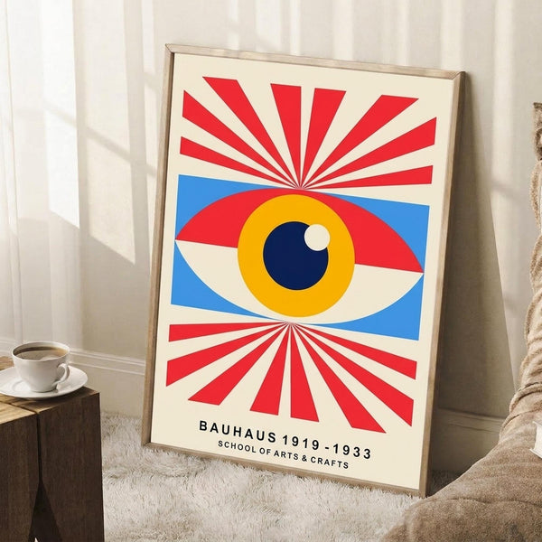 May include: Framed poster with abstract eye design featuring red and white radiating sunburst pattern, central eye with yellow iris and dark blue pupil surrounded by blue geometric shapes, displayed leaning against white wall next to wooden side table with white coffee cup and cozy knit throw blanket