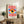 May include: Framed modernist poster featuring a large stylized eye with yellow iris, blue pupil, and white highlight, surrounded by red and white radiating sunburst rays above and below, with blue geometric shapes flanking the eye. The print sits on a rustic wooden console alongside books, a ceramic vase, and a potted fern, with a beige upholstered chair visible in a neutral-toned living room setting.