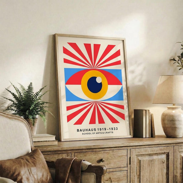 May include: Framed modernist poster featuring a large stylized eye with yellow iris, blue pupil, and white highlight, surrounded by red and white radiating sunburst rays above and below, with blue geometric shapes flanking the eye. The print sits on a rustic wooden console alongside books, a ceramic vase, and a potted fern, with a beige upholstered chair visible in a neutral-toned living room setting.