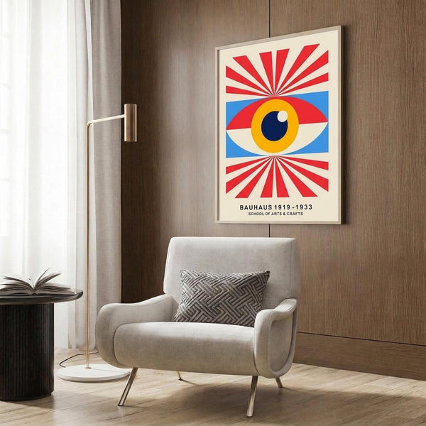 May include: Colorful Bauhaus-style poster with large eye design featuring red radiating lines, blue background, yellow and orange iris with dark blue pupil. Framed artwork displayed on wood-paneled wall above beige upholstered chair with geometric patterned pillow. Modern floor lamp with gold finish and open book on dark side table visible in minimalist interior setting.