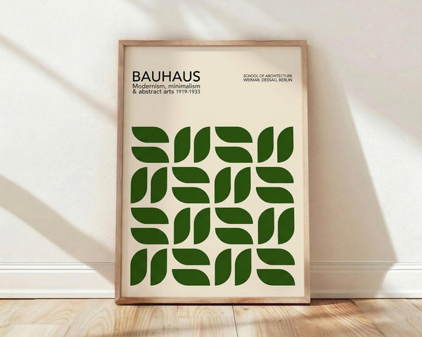 May include: Framed poster with cream background featuring geometric green leaf-like shapes arranged in a repeating pattern. Black text at top reads "BAUHAUS" with subtitle about modernism, minimalism and abstract arts 1919-1933, plus "SCHOOL OF ARCHITECTURE WEIMAR, DESSAU, BERLIN." Natural wood frame leaning against white wall on light wooden floor with soft shadows.