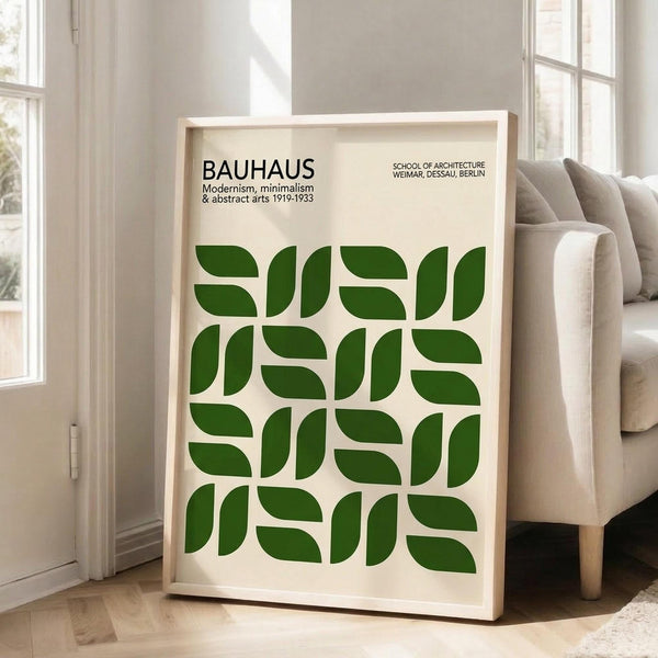 May include: Framed Bauhaus-inspired poster with cream background and geometric green leaf pattern in a 4x4 grid. Black text reads "BAUHAUS Modernism, minimalism & abstract arts 1919-1933" and "SCHOOL OF ARCHITECTURE WEIMAR, DESSAU, BERLIN." Light wood frame leans against white wall next to beige upholstered chair in bright, minimalist living room with large windows and hardwood floors.