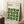 May include: Framed Bauhaus poster with green leaf pattern leaning against white wall corner. Light wooden frame contains cream background with repeated geometric green leaves arranged in grid pattern. Black text reads "BAUHAUS Modernism, minimalism & abstract arts 1919-1933" and "SCHOOL OF ARCHITECTURE WEIMAR, DESSAU, BERLIN." Coffee cup on wooden block nearby, beige throw blanket visible, natural window lighting.