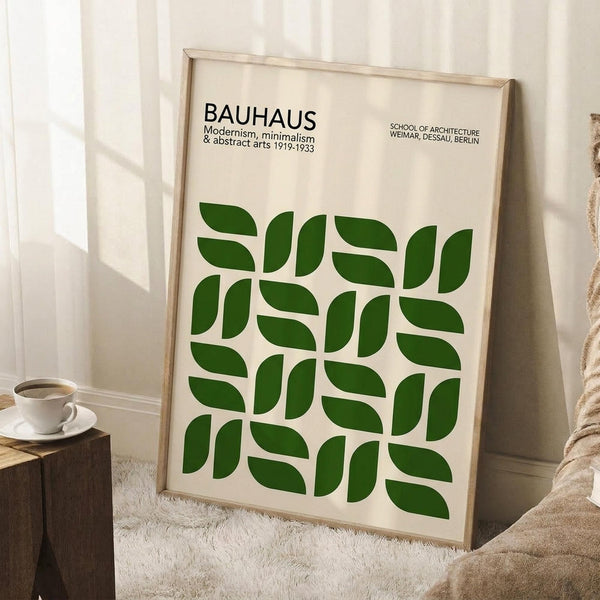 May include: Framed Bauhaus poster with green leaf pattern leaning against white wall corner. Light wooden frame contains cream background with repeated geometric green leaves arranged in grid pattern. Black text reads "BAUHAUS Modernism, minimalism & abstract arts 1919-1933" and "SCHOOL OF ARCHITECTURE WEIMAR, DESSAU, BERLIN." Coffee cup on wooden block nearby, beige throw blanket visible, natural window lighting.