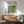 May include: Framed Bauhaus poster with green geometric leaf pattern hanging above mid-century modern wooden credenza. Mint green upholstered chair with wooden arms sits nearby, white ceramic table lamp on credenza. Warm sunlight creates diagonal shadows across white wall and wooden floor.