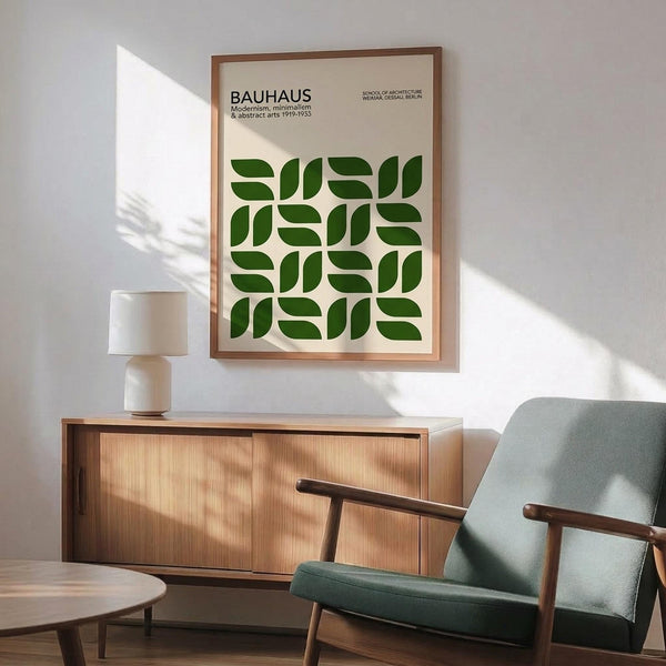 May include: Framed Bauhaus poster with green geometric leaf pattern hanging above mid-century modern wooden credenza. Mint green upholstered chair with wooden arms sits nearby, white ceramic table lamp on credenza. Warm sunlight creates diagonal shadows across white wall and wooden floor.