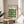 May include: Framed poster with green geometric leaf pattern displayed on wooden round side table next to beige upholstered chair. Poster features "BAUHAUS" text at top with repeating curved green shapes arranged in grid pattern on cream background. Natural lighting creates soft shadows on white wall behind, with plant leaves visible in foreground.
