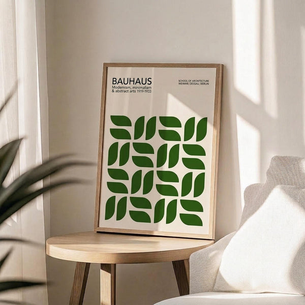 May include: Framed poster with green geometric leaf pattern displayed on wooden round side table next to beige upholstered chair. Poster features "BAUHAUS" text at top with repeating curved green shapes arranged in grid pattern on cream background. Natural lighting creates soft shadows on white wall behind, with plant leaves visible in foreground.