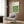 May include: A framed Bauhaus poster with green geometric leaf-like shapes arranged in a grid pattern on cream background, displayed on wood paneled wall above a beige upholstered armchair with patterned throw pillow. Modern brass floor lamp and round black side table with open book visible in warmly lit contemporary interior setting.