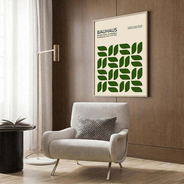 May include: A framed Bauhaus poster with green geometric leaf-like shapes arranged in a grid pattern on cream background, displayed on wood paneled wall above a beige upholstered armchair with patterned throw pillow. Modern brass floor lamp and round black side table with open book visible in warmly lit contemporary interior setting.